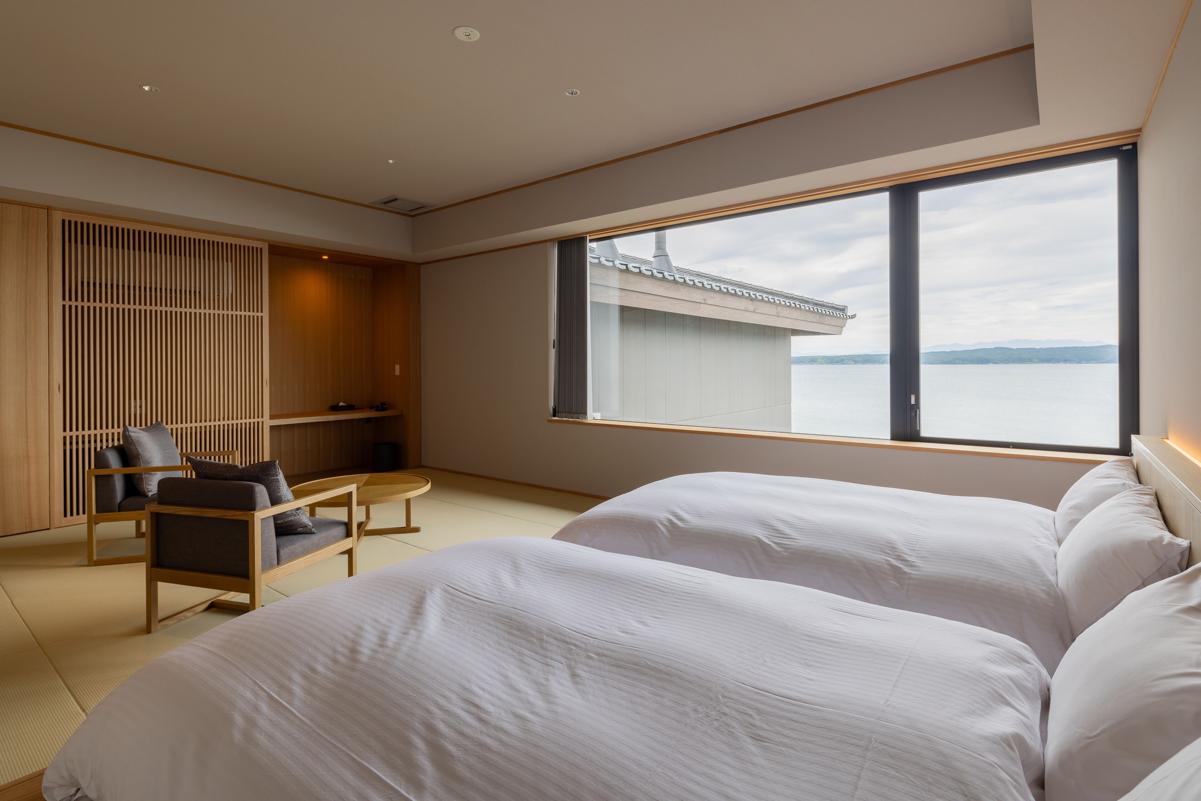 Quarto de hotel moderno com duas camas, uma cadeira de madeira, uma mesa pequena e uma janela grande com vista para um edifício e para a água.