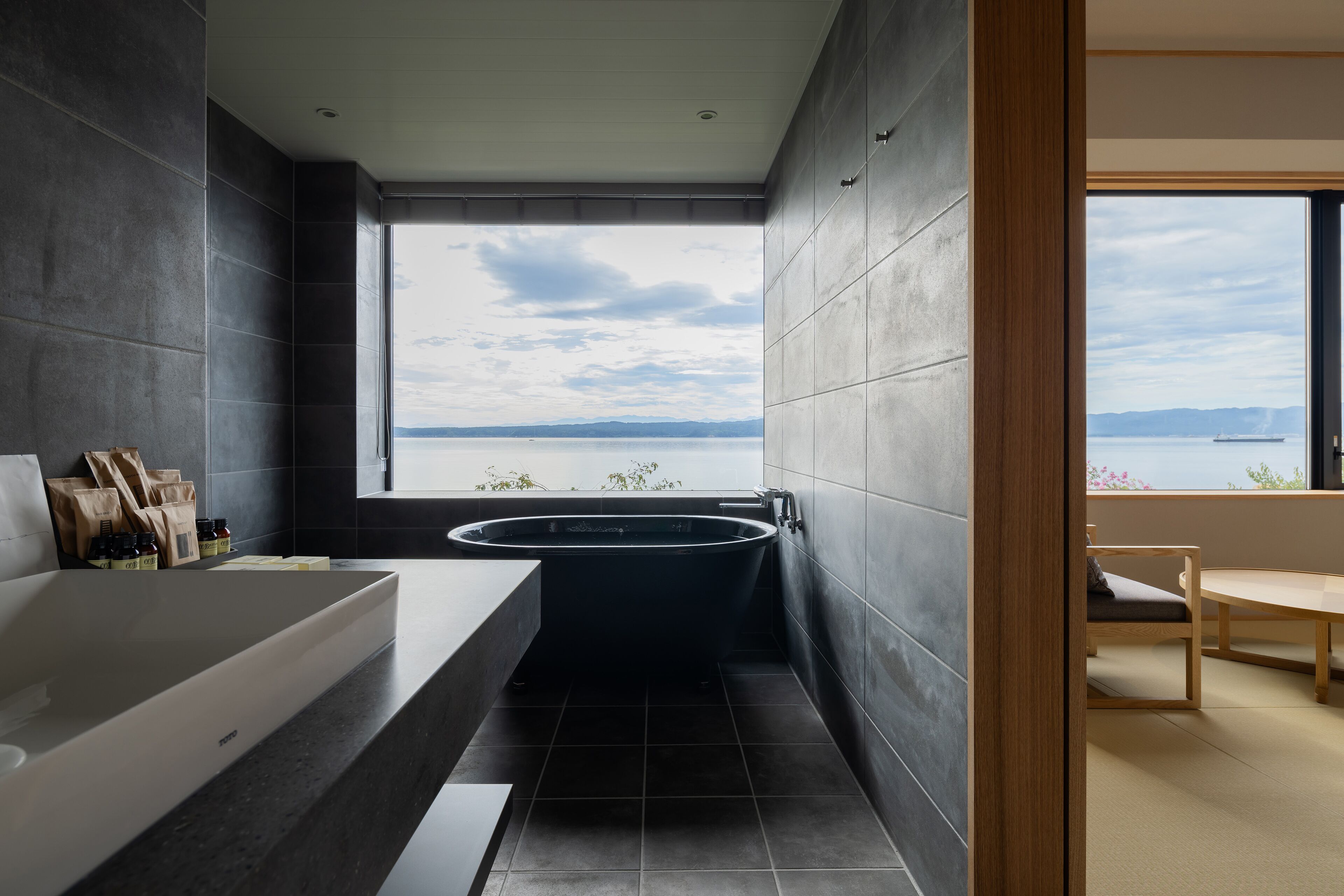 Un baño moderno con una bañera independiente, un amplio ventanal con vistas al mar y una silla de madera en un espacio luminoso.