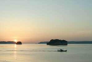 A calm body of water with a boat in the foreground and a large island in the background during sunset. - HITOTSU NOTOJIMA (Nanao)