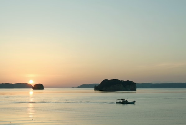 A calm body of water with a boat in the foreground and a large island in the background during sunset. - HITOTSU NOTOJIMA (Nanao)