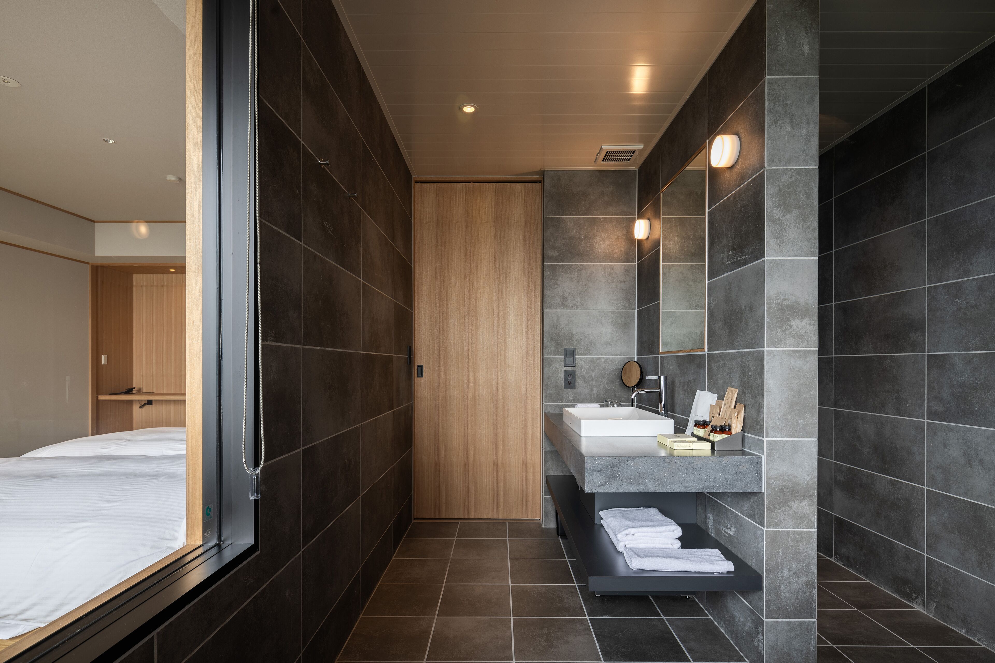 Un baño moderno con un espejo grande, baldosas oscuras, una puerta de madera y una cama empotrada.