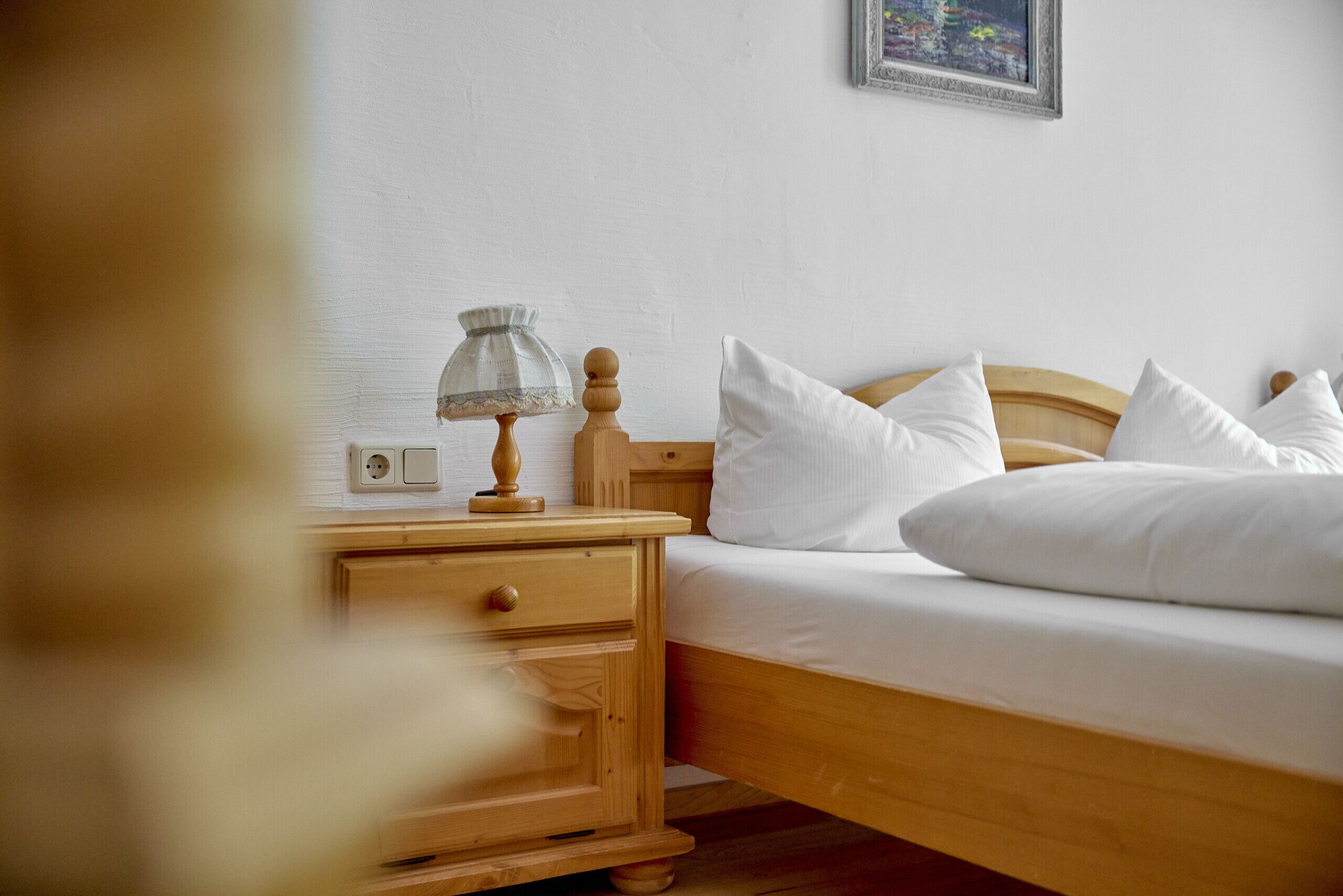 A neatly made bed with white linens and pillows, a wooden nightstand with a lamp, and a wall-mounted power outlet.