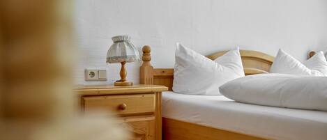 A neatly made bed with white linens and pillows, a wooden nightstand with a lamp, and a wall-mounted power outlet.