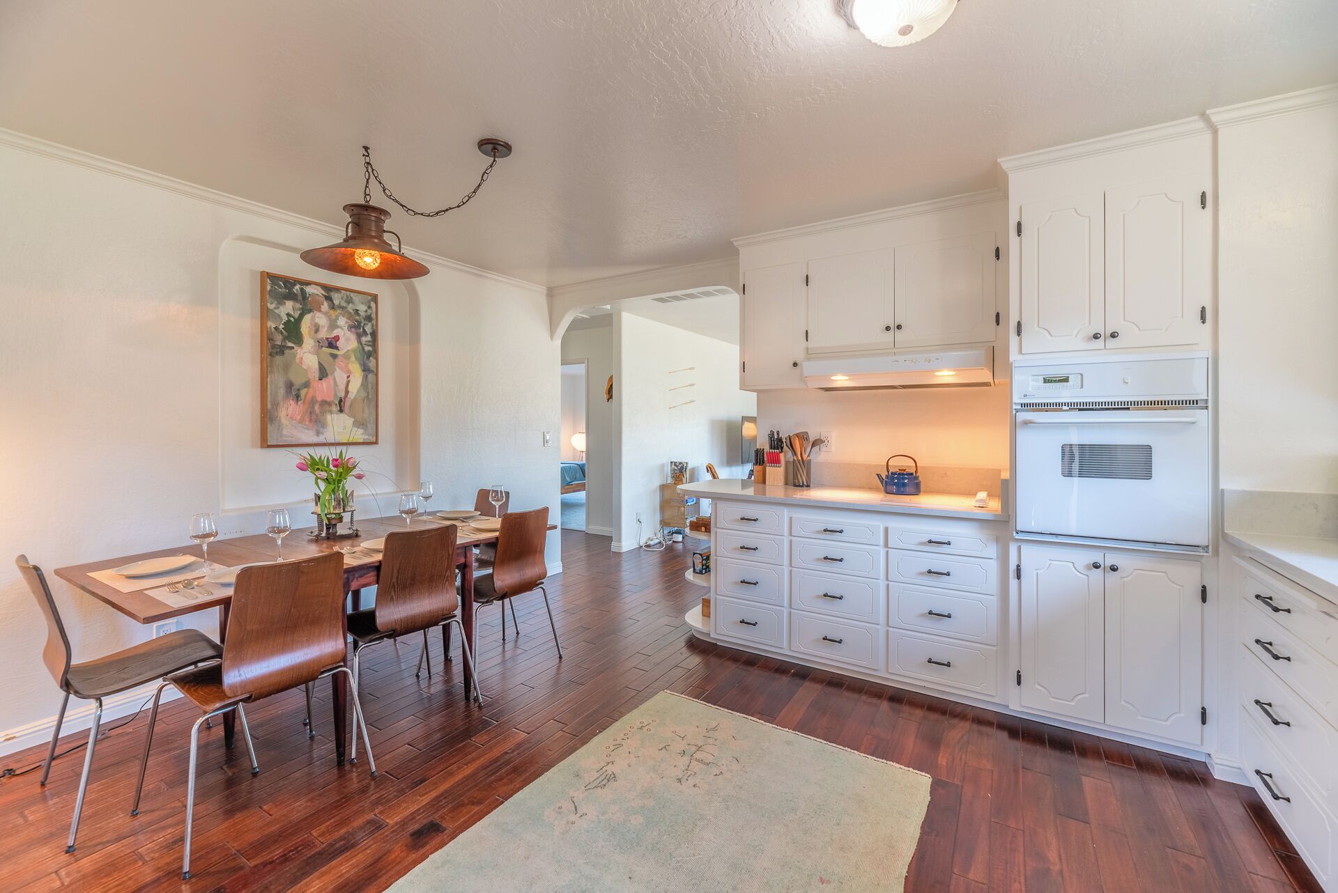 Un coin repas avec une table et des chaises en bois, un tableau au mur et des meubles de cuisine blancs dans la pièce adjacente.