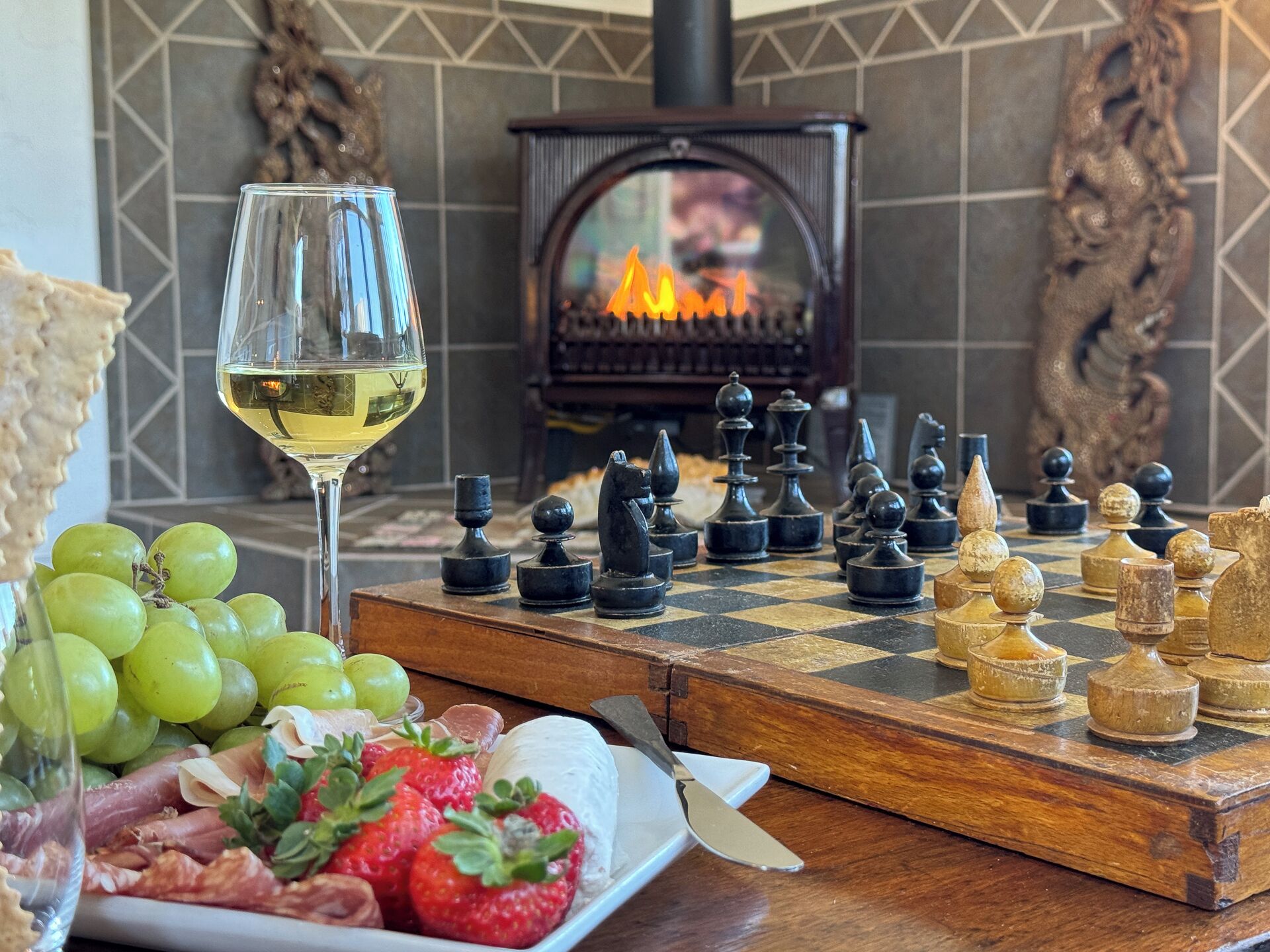 A chessboard with pieces, a glass of white wine, and a plate of food including grapes and strawberries.