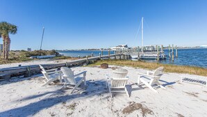 Un espace aménagé au bord de la plage, avec des chaises blanches et une table, une jetée en bois et des bateaux sur l’eau.