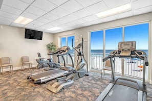 Een fitnessruimte met loopbanden, elliptische trainers en een hometrainer, grote ramen met uitzicht op het strand, en een flatscreen aan de muur.