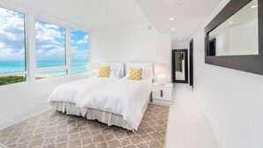 Une chambre moderne avec un grand lit, une literie blanche et deux oreillers jaunes. Elle est dotée d’une grande fenêtre offrant une vue sur l’océan et d’un plafond avec des spots encastrés.