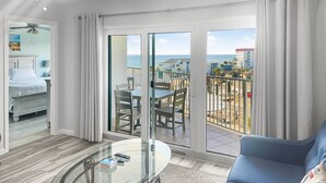 Una habitación de hotel moderna con balcón, una mesa de comedor de vidrio, un sofá azul y una cama con ropa blanca.