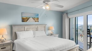 Un dormitorio con cama, mesitas de noche, ventilador de techo, un cuadro y un balcón con vistas.