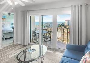 A modern hotel room with a balcony, a glass dining table, a blue sofa, and a bed with white bedding. - Seacrest 609 - Beachfront 2BR | Gulf View, Pool + Beach Service (Fort Walton Beach)