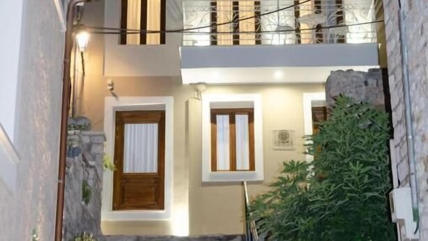 A two-story building with a stone staircase leading to the entrance, a balcony with metal railings, and a clock tower in the background. - Leone Suites (Nafpaktos)