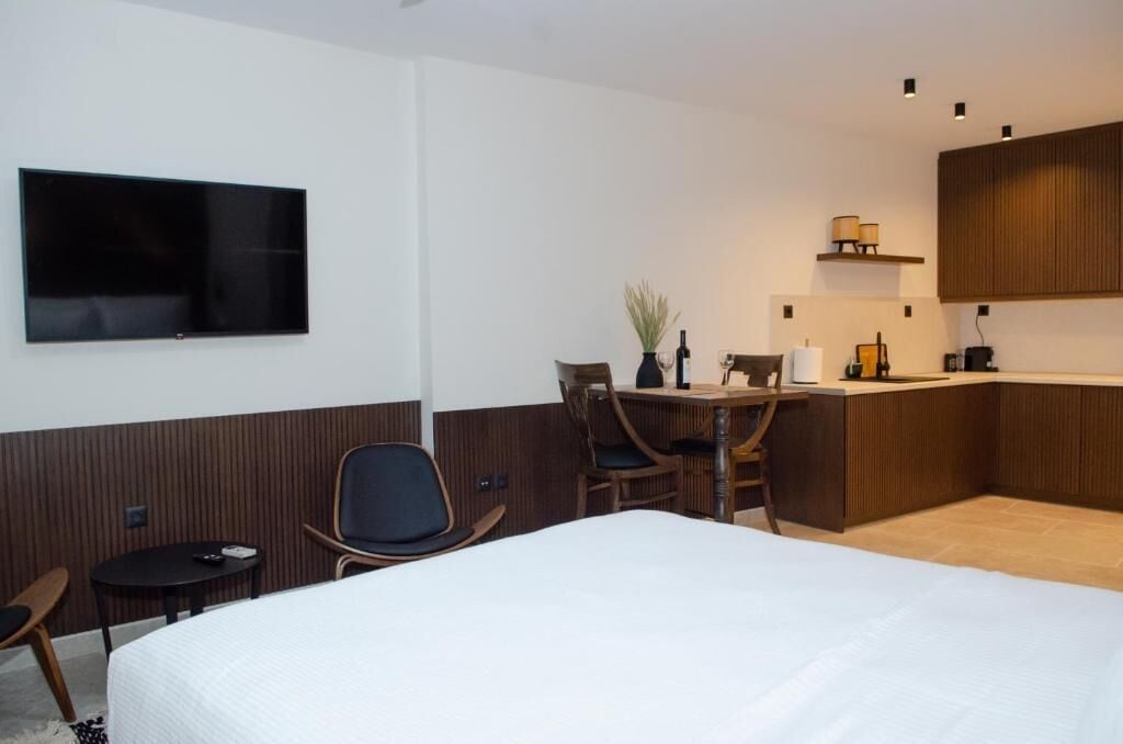 A hotel room with a bed, a TV mounted on the wall, a small table with a chair, a desk with a chair, and a kitchenette area.