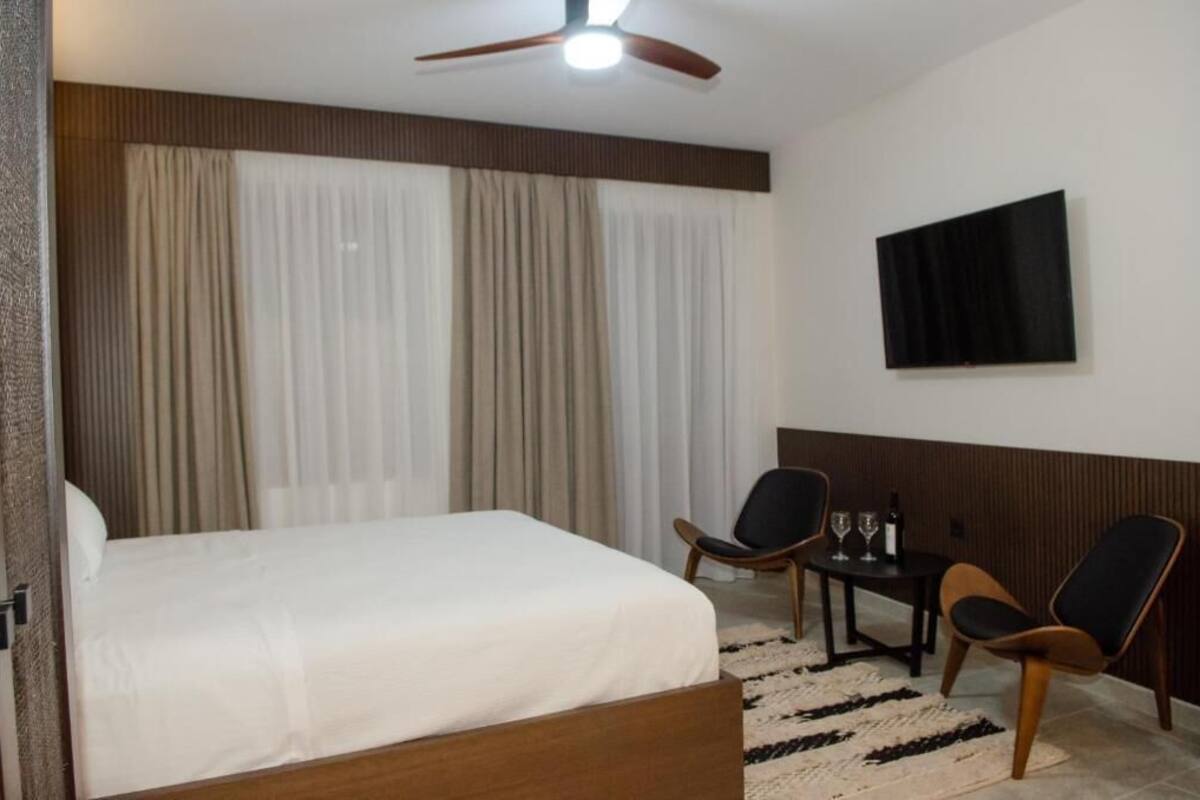 A hotel room with a bed, two chairs, a small table with glasses and a bottle, and a TV on the wall.
