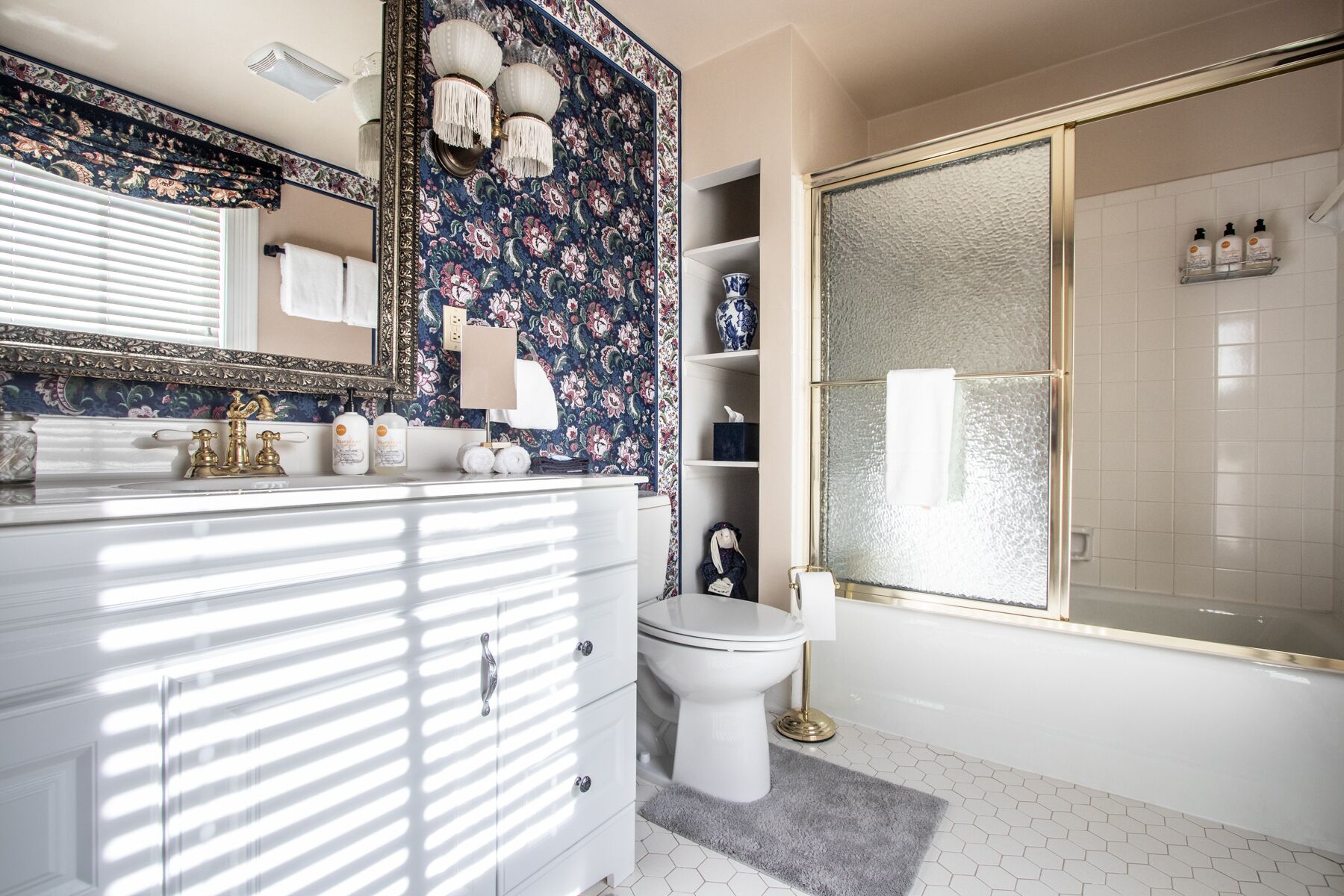 Un baño con tocador blanco, un espejo grande, un papel tapiz estampado con motivos florales, un inodoro y una ducha con puerta de cristal.