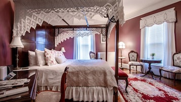 A four-poster bed with a lace canopy, a red and white patterned rug, and a wooden floor.