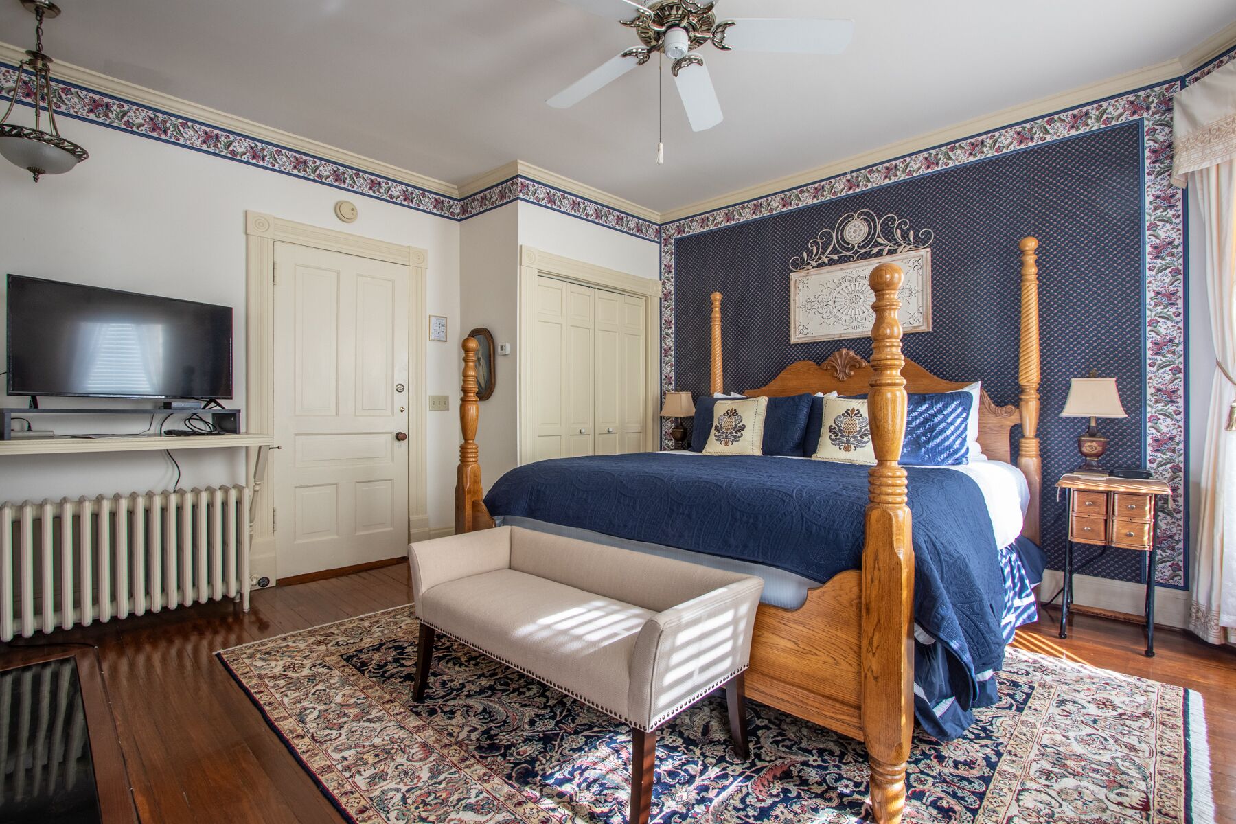 A bedroom with a four-poster bed, a television on a stand, a radiator, a bench, and a patterned rug.