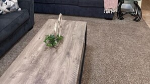 Ein Wohnzimmer mit einem dunkelgrauen Sofa, einem Holztisch mit Pflanze, einem Bild an der Wand, einer Lampe und einem kleinen Tisch ebenfalls mit einer Lampe.