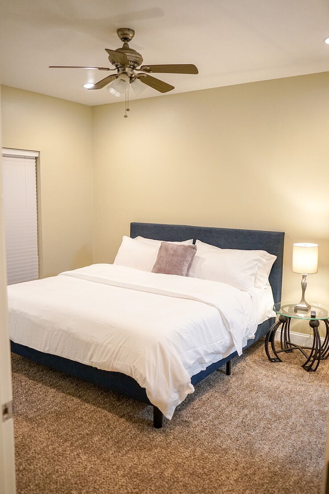 A bedroom with a bed, a ceiling fan, a bedside lamp, and a window with blinds.