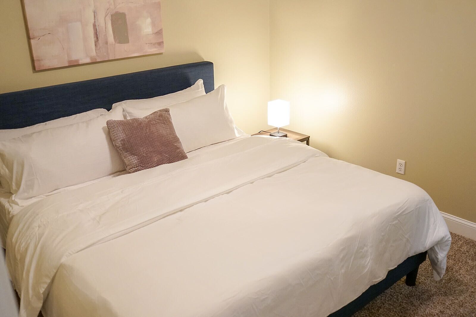 A neatly made bed with white linens and a dark headboard, a bedside lamp, and a plain wall with a single abstract painting.