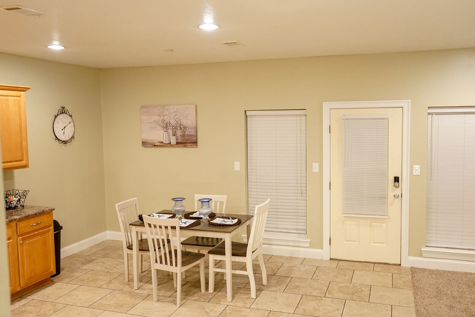 A dining area with a table set for four, surrounded by chairs, with a clock and artwork on the wall.