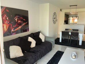 A modern living room with a black sofa, a dining area with a white table, and a kitchen in the background.