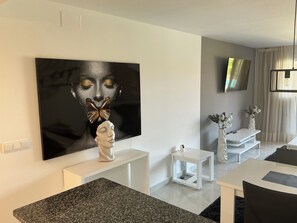Ein modernes Wohnzimmer mit einem großen abstrakten Gemälde, einem Flachbildfernseher, weißen Möbeln und einem Essbereich.