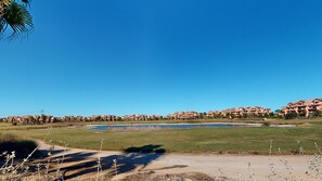 Ein Golfplatz mit blauem Himmel, einem Wasserhindernis und einer Reihe mehrstöckiger Gebäude im Hintergrund.