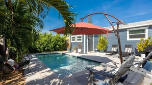 A swimming pool with lounge chairs and a red umbrella, surrounded by greenery and a house in the background.