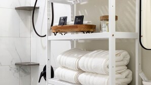 A bathroom with a white tiled wall, a white shelf with towels, and a shower area.
