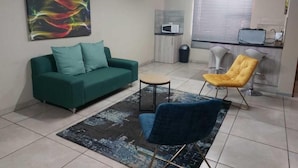 A living room with a green sofa, a blue chair, a yellow chair, a small round table, and a colorful abstract painting on the wall. - Wisteria Lane Guesthouse (Tsantsabane)