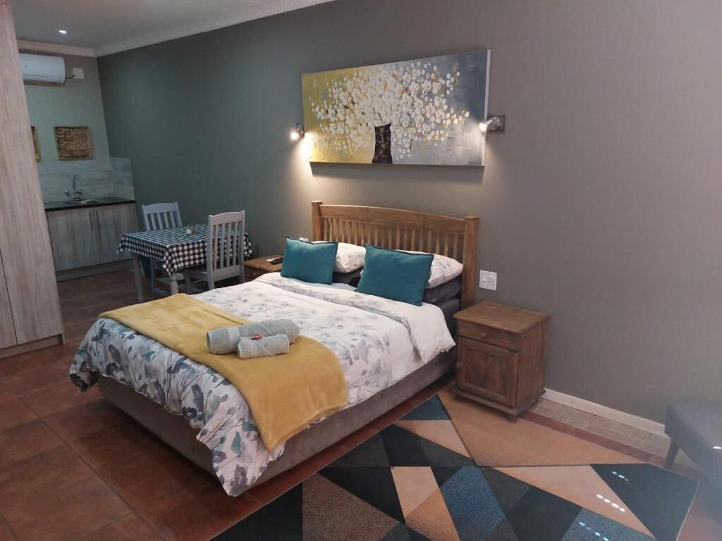 A bedroom with a bed, bedside tables, a painting, and a view of a kitchen area.