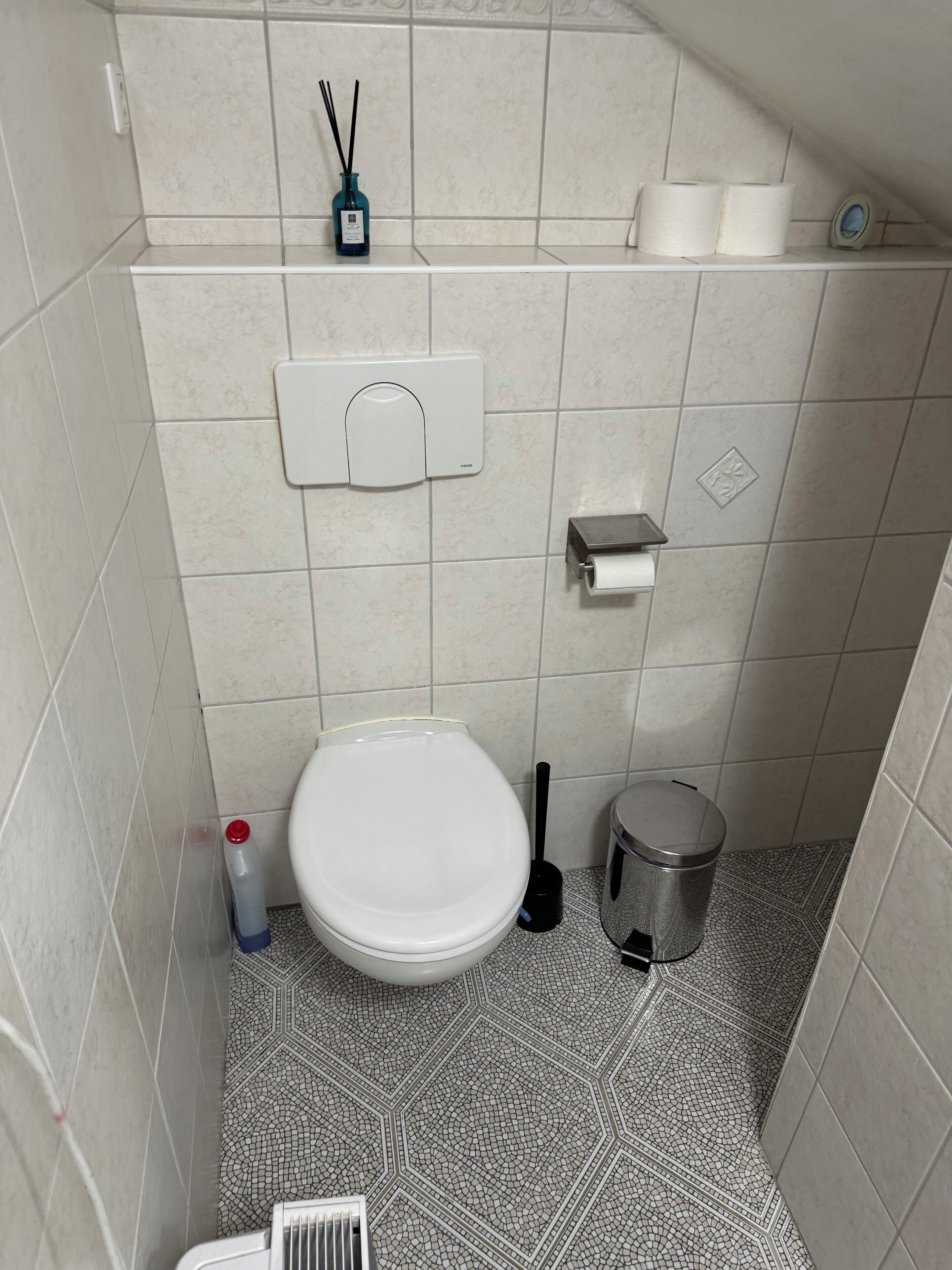 A small, tiled bathroom with a toilet, a bidet, a trash bin, a toilet paper holder, and a hand dryer.