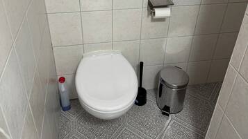 A small, tiled bathroom with a toilet, a bidet, a trash bin, a toilet paper holder, and a hand dryer.