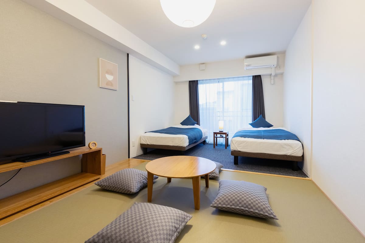 A hotel room with a TV, two beds, a small table, and futon seating.