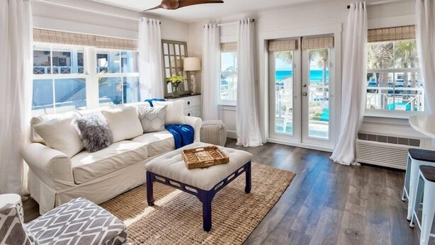 A living room with a sofa, ottoman, and a ceiling fan. There are large windows with curtains, a view of the outdoors, and a small table with a lamp. - Playa Bonita on 30A - Gulf View / Pickleball / Deeded Beach Access (Santa Rosa Beach)