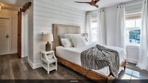 A bedroom with a wooden bed frame, white bedding, a nightstand with a lamp, a ceiling fan, and a view of the outdoors through a window with curtains. - Playa Bonita on 30A - Gulf View / Pickleball / Deeded Beach Access (Santa Rosa Beach)