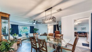 Un coin repas avec une table en verre et des chaises en bois, un ventilateur de plafond et une vue sur un salon avec un canapé et des plantes.