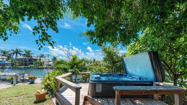 Une terrasse en bois avec un jacuzzi, entourée de plantes en pot et de palmiers, offrant une vue sur une étendue d’eau et une marina.