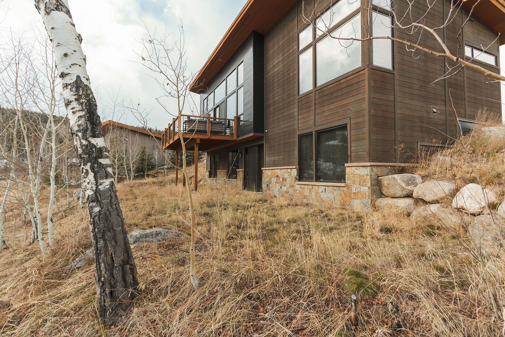 Une maison moderne à deux étages, avec des fondations en pierre, de grandes fenêtres et une terrasse en bois.