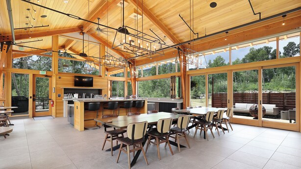 Un vaste espace de salle à manger avec une grande table et des chaises en bois, un coin cuisine avec un bar, et de grandes fenêtres offrant une vue sur l’extérieur.