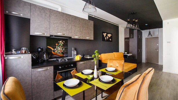 Une cuisine moderne avec des meubles gris, un plan de travail noir et un coin repas agrémenté de sets de table jaunes et de chaises orange.
