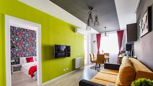 Un salon moderne avec un mur d’accent vert, un téléviseur à écran plat, un canapé, un coin repas avec des chaises et un coin cuisine avec une cuisinière.