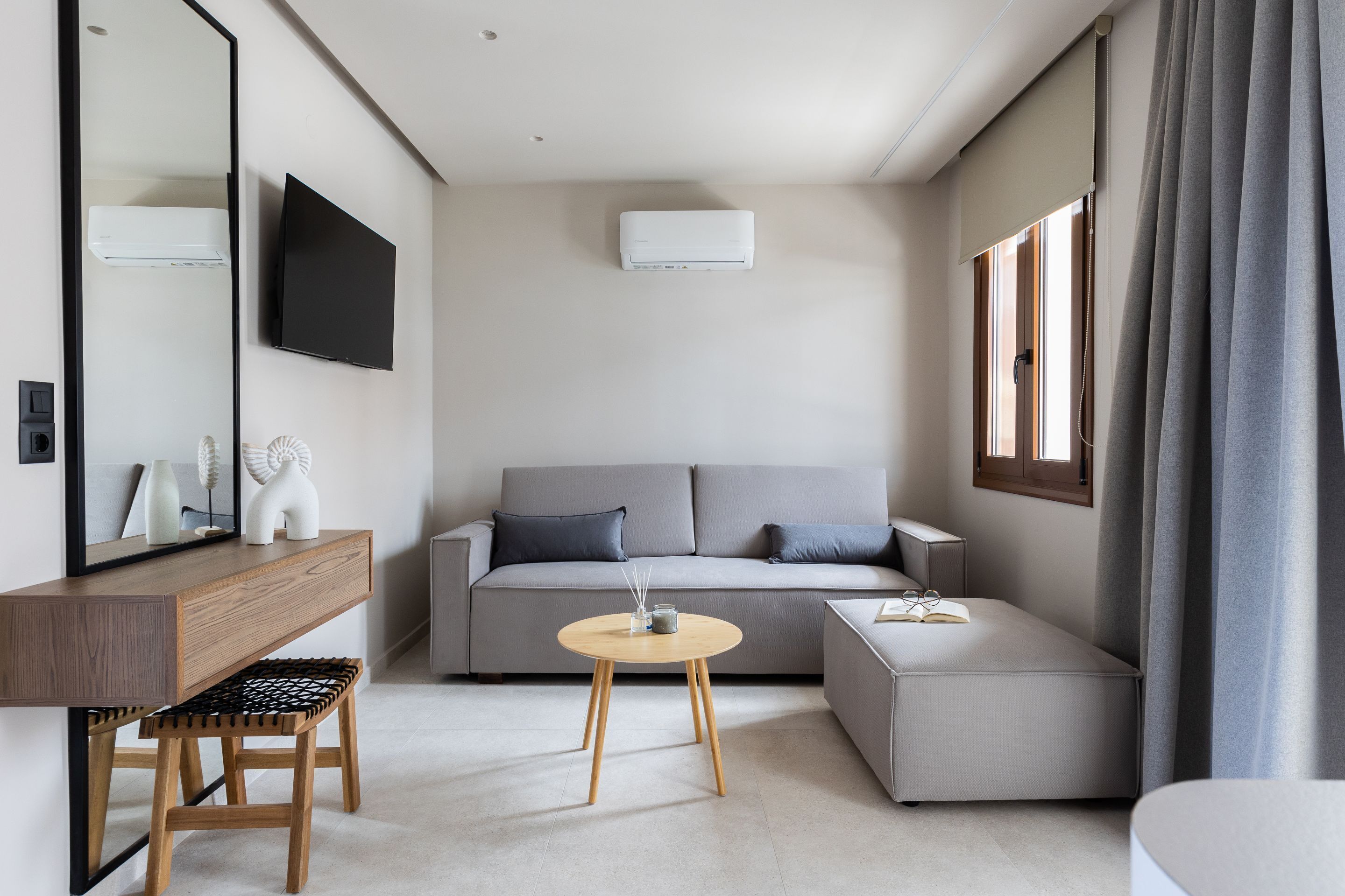 A modern, minimalist hotel room with a sofa, a small round table, a wooden bench, and a large mirror.