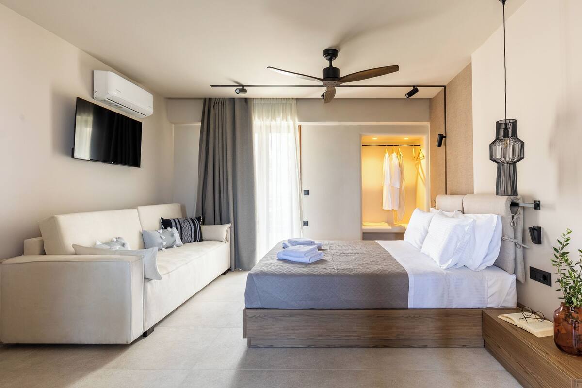 A modern hotel room with a bed, a sofa, a ceiling fan, an air conditioning unit, and a TV.
