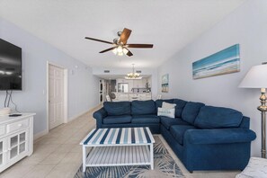 A living room with a blue sofa, a white coffee table, a ceiling fan, and a kitchen in the background.