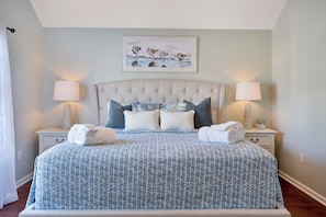 A bedroom with a tufted headboard, a patterned bedspread, two bedside lamps, and a painting of birds on the wall. - Z’s At The Sea | Surfside Beach 4BR Walk to Beach (Surfside Beach)