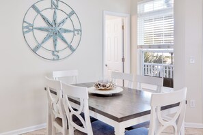 A dining area with a wooden table, white chairs, a decorative compass wall art, and a window with blinds.