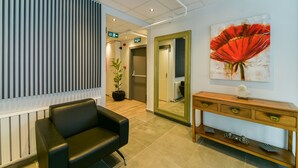 A reception area with a black leather chair, a wooden console table with drawers, a large mirror, and a red flower painting on the wall. - Rise Inn  (Istanbul)