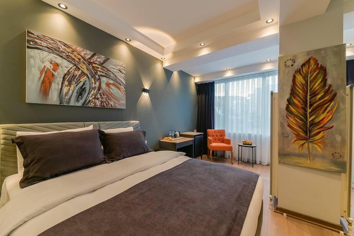 Moderni hotellihuone, jossa on suuri sänky, työpöytä ja tuoli. Seinillä on kaksi taulua ja ikkunassa on verhot.
